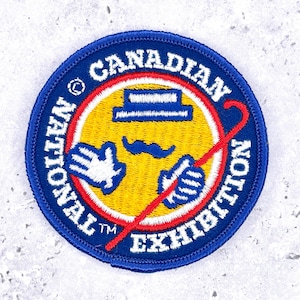 May include: Embroidered patch with the text "NATIONAL CANADIAN EXHIBITION" in a circular design. The patch features a yellow center with a cartoon face, a blue border, and a red cane crossing the center.