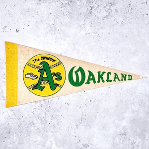 May include: Vintage pennant in a triangular shape, featuring the text "Oakland" in green, and a baseball graphic with the text "The Swingin' A's". The pennant has a yellow border and a cream-colored background.