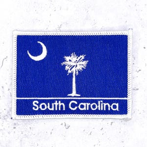 May include: A rectangular embroidered patch featuring the South Carolina state flag design. The patch has a deep blue background with a white crescent moon in the upper left corner, a white palmetto tree in the center, and the text South Carolina.