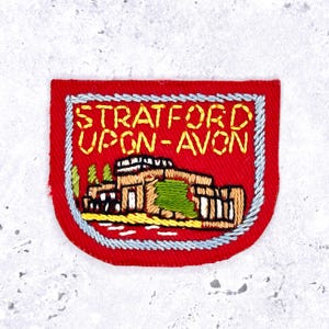 May include: A red embroidered patch with a light blue border featuring the yellow text STRATFORD UPON-AVON above a depiction of a building with brown walls, a white roof, and green accents, all rendered in detailed thread work on a red fabric base.