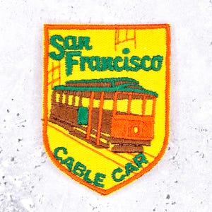 Op de afbeelding: Geborduurde patch met een ontwerp van een San Francisco kabeltram. De schildvormige patch heeft een gele achtergrond en een oranje rand. De tekst "San Francisco Cable Car" is groen.