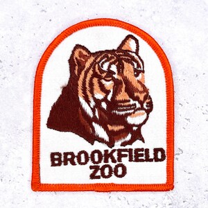Op de afbeelding: Geborduurde patch met een tijgerkop in bruin en oranje op een witte achtergrond. De patch heeft een oranje rand en de woorden "BROOKFIELD ZOO" zijn in het zwart weergegeven. Een souvenirartikel.