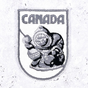 Könnte beinhalten: Gestickter Aufnäher mit dem Wort "CANADA" oben. Darunter eine detaillierte Illustration einer Inuit-Figur in traditioneller Kleidung. Der Aufnäher hat einen weißen Hintergrund mit einem grauen Rand und die Figur ist in Grautönen gehalten.