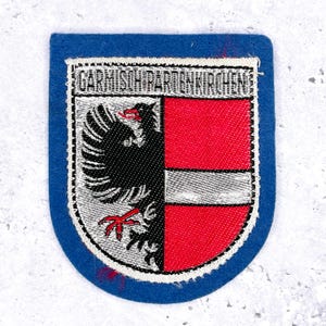 May include: A blue patch with a shield design. The shield is divided into halves: one black with a bird, the other red. The text "GARMISCH-PARTENKIRCHEN" is at the top.