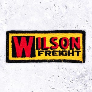 Op de afbeelding: Geborduurde patch met de tekst "WILSON FREIGHT" in rood en zwart op een gele achtergrond. De patch heeft een zwarte rand en een rechthoekige vorm.