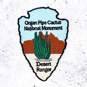 May include: Embroidered patch in the shape of an arrowhead, featuring the text "Organ Pipe Cactus National Monument" and "Desert Ranger." The design includes a light blue sky, brown mountains, a green cactus, and a tan desert floor.