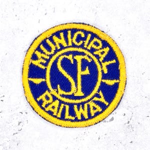May include: A circular embroidered patch featuring a deep blue background with a bright yellow border and yellow text. The center displays the letters SF in a bold font, while the text MUNICIPAL RAILWAY is stitched along the circular edge.