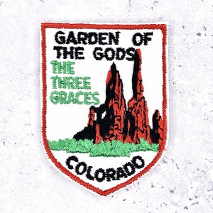 Op de afbeelding: Geborduurde patch met de tekst "GARDEN OF THE GODS THE THREE GRACES COLORADO". De patch heeft een witte achtergrond en een rode rand. Het ontwerp bevat rode rotsformaties, groen gras en zwarte tekst.