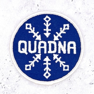 Könnte beinhalten: Ein kreisförmiger bestickter Aufnäher mit tiefblauem Hintergrund und weißem Rand. Das Wort QUADNA ist in weißen Großbuchstaben in die Mitte gestickt, umgeben von einem weißen, schneeflockenartigen geometrischen Design mit sechs Spitzen.
