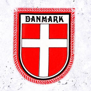May include: A shield-shaped embroidered patch featuring the red and white flag of Denmark. The word DANMARK is printed in black capital letters at the top. The patch has a thick black border and a red decorative stitched edge around the perimeter.