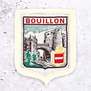 Può includere: Una toppa a forma di scudo color crema con la scritta "BOUILLON" in lettere rosse. La toppa presenta un'immagine dettagliata di un castello e uno stemma. Il bordo è color crema con un motivo a puntini.