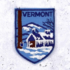 Op de afbeelding: Geborduurde patch met een winterlandschap met een met sneeuw bedekt huis en bergen onder het woord "VERMONT" in blauw. De patch heeft een schildvorm met een blauwe rand en een bruine boomstam.