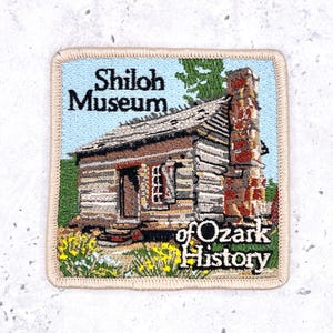 May include: A square embroidered patch featuring a log cabin with a stone chimney, green trees, and yellow flowers. The text Shiloh Museum of Ozark History is stitched in black and white lettering against a light blue sky and green landscape background.