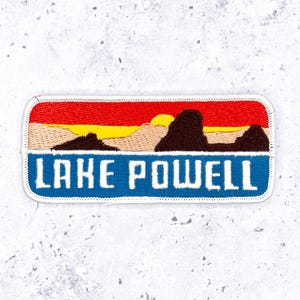 May include: A rectangular embroidered patch featuring a landscape scene of dark brown rock formations against a bright yellow sun and a vibrant red sky. Below the landscape, a blue section displays the white text LAKE POWELL in block letters.