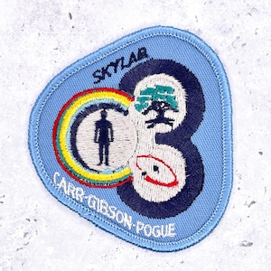 May include: A triangular embroidered patch with a light blue background. It features the text SKYLAB and CARR-GIBSON-POGUE in black. The design includes a central figure inside a rainbow circle, a dark blue number 3, and small graphic icons.