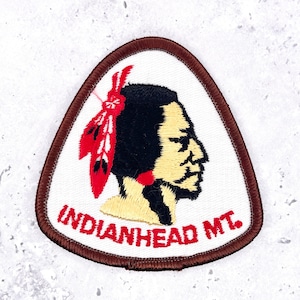 May include: A triangular embroidered patch with a brown border on a white background. It features a profile illustration of a person with black hair, a red feather, and tan skin. The red text "INDIANHEAD MT." is stitched along the bottom.