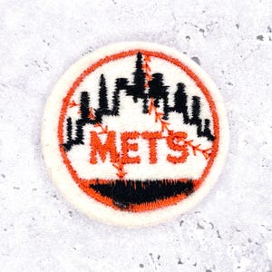 Może przedstawiać: Okrągła naszywka z logo New York Mets. Projekt zawiera pomarańczowy napis "METS", czarną panoramę miasta i pomarańczowy kontur szwu baseballowego. Naszywka jest na białym tle.