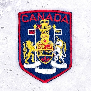 May include: A shield-shaped embroidered patch with a dark blue background and a red border. The word CANADA is stitched in red at the top. The centre features a yellow lion and a white unicorn flanking a central crest with red and yellow details.