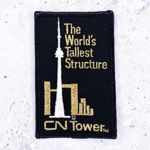 May include: Black rectangular patch featuring the CN Tower in white and gold embroidery. The text "The World's Tallest Structure" is above the tower, and "CN Tower" is below. The patch has a black border.