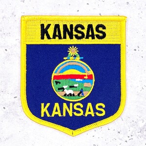 May include: A shield-shaped patch with a yellow border and the word "KANSAS" in black at the top and bottom. The center is blue with a circular emblem featuring a landscape scene.