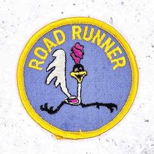 May include: Embroidered patch featuring the Road Runner cartoon character. The patch has a blue background with the words "ROAD RUNNER" in yellow. The Road Runner is depicted in white, black, pink, and yellow.