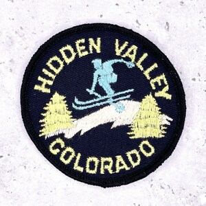 May include: A round, navy blue patch with the words "HIDDEN VALLEY COLORADO" in yellow lettering. The patch features a skier in light blue and two yellow pine trees. The skier is in motion, skiing down a slope.