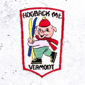 May include: A shield-shaped embroidered patch with a red border featuring a cartoon pig wearing a red hat, a red scarf, a light pink sweater, and green trousers. The pig holds yellow and blue skis. Text reads HOGBACK Mt. and VERMONT.