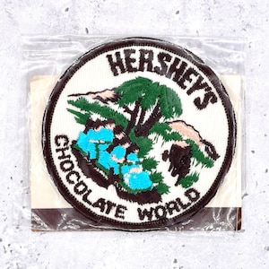 May include: Round embroidered patch featuring the words "Hershey's Chocolate World" in white lettering on a white background. The patch has a brown border and depicts a landscape with trees and water in shades of green and blue.