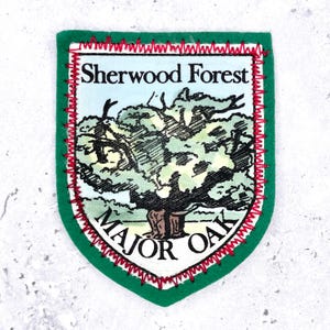 Op de afbeelding: Een schildvormige stoffen patch met een groene rand en rode zigzagstiksels. Het midden toont een gedrukte illustratie van een grote boom met de tekst Sherwood Forest erboven en Major Oak eronder in zwarte letters op een lichte achtergrond.