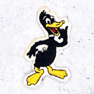 May include: Embroidered patch featuring a cartoon duck in black, yellow, and red. The duck is depicted with a white outline, a yellow beak and feet, and a red tongue. The patch is likely for clothing or accessories.