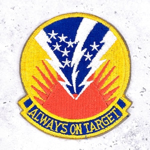 May include: Embroidered patch with a yellow border, featuring a blue and white design with stars and lightning bolts. The patch has a red base and the text "ALWAYS ON TARGET" in blue.