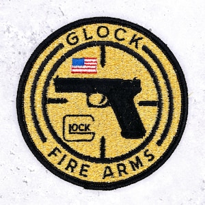 May include: A circular embroidered patch with a gold background and black border. It features a black handgun silhouette, a small American flag, and the text GLOCK FIRE ARMS. The design includes a crosshair graphic centered behind the handgun.