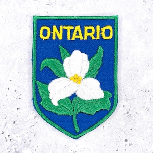 May include: A shield-shaped embroidered patch featuring a blue background with a green border. The word ONTARIO is stitched in yellow capital letters at the top. A white trillium flower with a yellow center and green leaves is centered.
