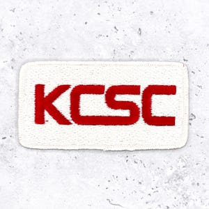 May include: A rectangular white patch with the letters "KCSC" embroidered in red. The letters are bold and stylized, with a slightly fuzzy texture. The patch is likely designed to be sewn or ironed onto clothing or other fabric items.