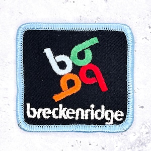 May include: A square patch with a black background and a light blue border. The patch features the word "breckenridge" in white, and colorful stylized letters. The letters are white, green, orange, and red.