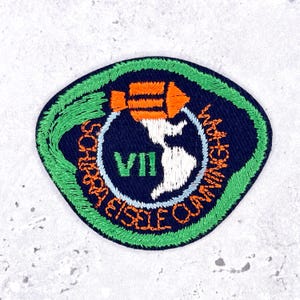 Pode incluir: Um emblema bordado circular com fundo azul escuro e borda verde brilhante. Apresenta um foguete laranja, uma silhueta branca das Américas, o numeral romano VII em verde e o texto laranja Schirra Eisele Cunningham.