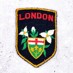 May include: A black embroidered patch with the word "LONDON" in red. The patch features white flowers, green leaves, and a shield with a red cross and yellow and green accents. The border is yellow.