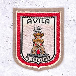 May include: A rectangular patch with a shield design featuring the text "AVILA" at the top and "AVILA DEL REY" at the bottom. The shield depicts a castle with a knight on top. The patch has a red border and a tan background.