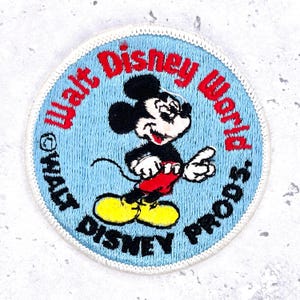 May include: Round embroidered patch featuring Mickey Mouse with the text "Walt Disney World PRODS." in red and blue. Mickey is in a classic pose, wearing red shorts and yellow shoes, set against a light blue background.