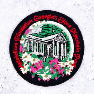 May include: A circular embroidered patch featuring a white building with columns, surrounded by green foliage and pink and white flowers. The black border contains the red embroidered text: Antebellum Plantation Georgia's Stone Mountain Park.