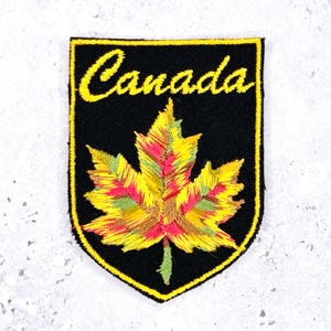May include: Embroidered patch featuring a colorful maple leaf in shades of yellow, red, and green, set against a black shield-shaped background. The word "Canada" is stitched in yellow script across the top.