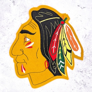May include: An embroidered patch featuring the profile of a person with yellow skin, black hair, and a red earring. The person wears a headdress with red, green, yellow, and orange feathers. There are red and white markings on the cheek and chin.