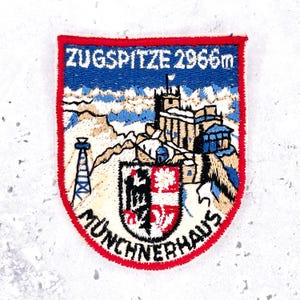 May include: A shield-shaped embroidered patch featuring a mountain landscape with the text ZUGSPITZE 2966m and MÜNCHNERHAUS. The design includes a building, a tower, and a coat of arms in red, white, blue, and black, all bordered by a red edge.