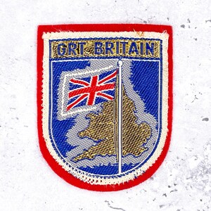May include: Embroidered patch with a red border and the text "GRI BRITAIN". The patch features a blue background with a gold-colored map of Great Britain and a Union Jack flag.