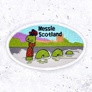May include: An embroidered patch featuring the Loch Ness Monster, Nessie, in a Scottish landscape. The patch has a white border and includes the text "Nessie Scotland". Nessie is green and wearing a red and green outfit.