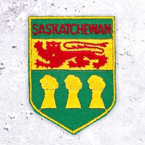 May include: Embroidered patch featuring the word "SASKATCHEWAN" above a red lion and three yellow wheat sheaves. The patch has a green border and yellow accents, resembling a provincial emblem.