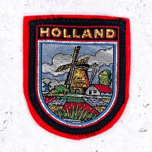 Può includere: Toppa ricamata con bordo rosso e finiture nere. La toppa presenta la scritta "HOLLAND" in lettere dorate sopra una scena dettagliata di un mulino a vento, campi e una casa con il tetto rosso.