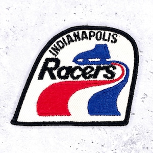 Może przedstawiać: Haftowana naszywka z napisem "Indianapolis Racers" w kolorze czarnym, ze stylizowanym niebiesko-czerwonym wzorem. Naszywka ma białe tło i czarną obwódkę. Prawdopodobnie naszywka drużyny sportowej lub pamiątka.