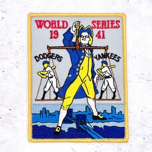 May include: Embroidered patch featuring a cartoon illustration of a man in colonial attire holding scales, with baseball players and the text "WORLD SERIES 1941 DODGERS YANKEES". The patch has a gold border and a blue and yellow color scheme.