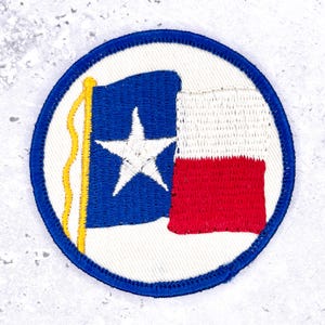 Peut inclure: Un écusson brodé circulaire sur fond blanc avec une bordure bleue. Le motif représente un mât jaune avec un drapeau composé d'une bande verticale bleue ornée d'une étoile blanche, suivie d'une bande horizontale blanche et d'une bande rouge.